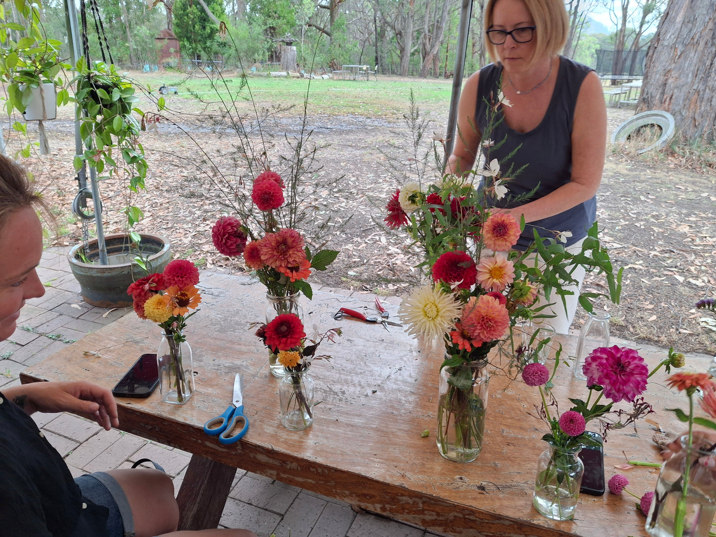 Two women arranging flowers at a wooden table outdoors.